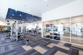 This fitness center is organized across a single level and designed to support a comprehensive range of workout activities. The space is divided into clearly defined zones, beginning with a bright entry area featuring a bold, geometric multicolor mural bearing the "Eisley" branding. This welcoming entrance includes built-in white shelving, a beverage cooler, and direct glass door access to the outdoor grounds, where palm trees and landscaped areas are visible.

The main workout floor is generously proportioned, finished with a patterned modular carpet in neutral tones of blue, gray, and tan that adds visual structure to the open layout. A dedicated strength training zone houses cable machines, multi-station cable systems, free weight benches, and a full dumbbell rack, all positioned beneath a distinctive navy blue tray ceiling fitted with recessed lighting and ceiling fans.

A separate glass-enclosed studio provides space for group fitness or stretching activities, with mirrored walls, organized equipment storage, and ample natural light. The cardio zone features treadmills and stationary bikes arranged along large sliding glass windows that frame views of the outdoor courtyard. Wall-mounted televisions and full-length mirrors throughout enhance both functionality and the sense of spaciousness.
