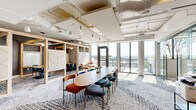 This contemporary shared workspace offers a well-appointed single-floor layout designed to support a variety of collaborative and focused work styles. The open central area features a large oval conference table surrounded by a curated mix of teal, navy, and rust-colored upholstered chairs, anchored by a patterned, multi-tonal carpet that adds visual texture throughout. Pendant globe lighting descends from exposed ceilings where white-painted ductwork and structural elements lend an industrial-modern aesthetic.

Natural light floods the interior through floor-to-ceiling glass walls and sliding doors that open directly onto an outdoor terrace, offering sweeping views of the surrounding landscape. Light wood open shelving units serve as stylish room dividers, creating semi-private lounge alcoves furnished with intimate seating arrangements ideal for small group discussions.

A glass-enclosed private meeting room provides a quiet, enclosed option for focused work or confidential conversations. The entry corridor features a vibrant teal accent wall framing a colorful cityscape artwork, a sleek light wood credenza, and warm hardwood flooring — setting an inviting, professional tone from the moment of arrival. The overall design balances openness with purpose-driven zones, making this a highly functional and visually engaging workspace.