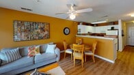 Located at 413 Sunchase Blvd in Farmville, Virginia, this single-floor residence offers a well-organized layout designed for comfortable everyday living. The living area makes an immediate impression with its bold golden-yellow accent wall, warm hardwood-style flooring, and a ceiling fan overhead. A sliding glass door floods the space with natural light and provides access to an outdoor area. The open flow connects seamlessly to a combined dining space, where a round wooden table sits adjacent to a peninsula-style kitchen. The kitchen features white cabinetry, light countertops, a double sink, a dishwasher, and a range, all set against a coordinating amber-toned wall. Multiple bedrooms are found throughout the unit, each fitted with ceiling fans, carpeted floors, and windows with blinds that allow for natural light and privacy. The bedrooms vary in character while maintaining a consistent, neutral palette suited for a range of uses. Two full bathrooms serve the residence, both finished with white vanity cabinetry, large mirrors with overhead lighting, tile flooring, and tub-shower combinations, offering clean and functional design throughout.