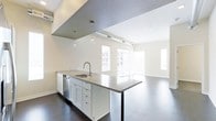 This contemporary single-floor space showcases a thoughtfully designed open-concept layout that seamlessly blends the kitchen and living areas. The kitchen features a large central island with a dark countertop, an undermount sink with a modern faucet, white shaker-style cabinetry, and a stainless steel dishwasher. Pendant lighting hangs above the island, adding a refined touch to the space. The ceiling transitions from a dark, textured surface over the kitchen to a bright white finish in the living area, creating a distinct yet cohesive visual separation between zones. Exposed ductwork and industrial-style track lighting add an urban, loft-inspired character to the space. The living area is bathed in natural light, courtesy of large windows with horizontal blinds and a narrow sidelight window that further brightens the room. Polished concrete floors run throughout, offering a sleek, low-maintenance surface that complements the modern aesthetic. The overall ambiance is clean, minimalist, and functional, making this space well-suited for those who appreciate contemporary design and open, airy interiors.