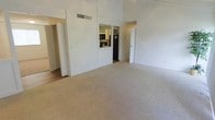This single-floor space is defined by its notably high vaulted ceilings, which lend a sense of openness and airiness to the main living area. The expansive living room is finished with plush, neutral-toned wall-to-wall carpeting that contributes to a clean, understated aesthetic throughout. Bright white walls reflect available light, enhancing the overall sense of space.

A pass-through window connects the living area to the kitchen, offering a glimpse of dark appliances, including a black refrigerator and microwave, set against light cabinetry. This architectural detail maintains visual connectivity between the spaces while preserving a degree of separation. The kitchen layout appears functional and efficiently organized.

Multiple doorways are visible from the main living area, suggesting access to additional rooms and hallways branching off in different directions. A dedicated HVAC vent is positioned near the ceiling, indicating a central climate control system. The overall layout is straightforward and versatile, suitable for a variety of furniture arrangements. The neutral finishes and open design provide a practical, move-in-ready foundation.