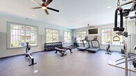 This fitness center offers a generously sized, single-room workout space designed to accommodate a comprehensive range of exercise equipment and training styles. The room features a clean, neutral color palette with soft blue-gray walls accented by board-and-batten wainscoting, creating a polished and inviting atmosphere. Large, multi-pane windows line multiple walls, flooding the space with abundant natural light and offering views of the surrounding greenery. Two ceiling fans provide consistent airflow throughout the room, while recessed lighting supplements illumination for evening use. A wall-mounted flat-screen television is positioned for easy viewing during workouts. The flooring consists of durable, dark rubberized material well-suited for athletic use. Equipment includes two treadmills, an elliptical trainer, a stationary bike, a rowing ergometer, and a multi-station cable machine, offering a full suite of cardio and strength-training options. A tiered dumbbell rack stocked with a wide range of free weights, along with an adjustable workout bench, rounds out the strength training area. A large wall mirror enhances the sense of space and allows for form checks during exercise. The overall ambiance is bright, functional, and well-maintained, making it an effective dedicated fitness facility.