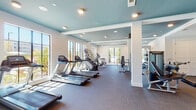 Located at 2620 Mall of Georgia Blvd in Buford, Georgia, this well-appointed fitness center offers a comprehensive workout environment designed for both cardio and strength training. The single-floor layout is thoughtfully organized into distinct functional zones, allowing users to move seamlessly between equipment types.

The cardio area is a standout feature, lined with multiple treadmills and elliptical machines positioned along large windows that invite abundant natural light and outdoor views. The bright, open feel of this zone is further enhanced by the distinctive teal-painted ceiling, which adds a vibrant yet calming visual contrast to the clean white walls.

The strength training section includes a well-stocked free weights area featuring tiered dumbbell racks with a full range of weights, an adjustable workout bench, and a large gold-framed mirror that visually expands the space and allows for form monitoring. A flat-screen television is mounted nearby for added entertainment during workouts. Additional cable and resistance machines round out the strength equipment offerings, providing options for a full-body training routine.

The facility features carpet flooring throughout, recessed lighting, and structural columns that define the different workout zones without fully enclosing them. Entry is through glass double doors, and the space is equipped with safety essentials including a fire extinguisher and emergency exit signage. The overall ambiance is clean, modern, and welcoming.