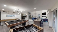 This single-level clubroom is a thoughtfully designed communal space that seamlessly blends entertainment, relaxation, and social gathering areas into one cohesive environment. The game zone anchors one end of the space, housing a full-size pool table and foosball table beneath a contemporary globe chandelier, setting a lively tone upon entry.

Adjacent to the game area, a fully equipped kitchen serves as a central hub for gatherings. Polished marble tile flooring leads to a substantial marble-wrapped island with bar seating, illuminated by elegant pendant lights. Dark cabinetry contrasts dramatically against a floor-to-ceiling gold hexagonal mirror tile backsplash, adding a glamorous, reflective quality to the space.

A dining area with a concrete-style table and upholstered seating transitions into the lounge zone, which features tufted gray velvet sofas arranged around a sleek linear fireplace topped by a wall-mounted television. Soaring double-height ceilings amplify the sense of volume in this area, while large sliding glass doors open directly to an outdoor patio, flooding the interior with natural light and offering seamless indoor-outdoor connectivity.

Curated wall art, metallic sculptural installations, and layered area rugs throughout contribute to the space's polished, contemporary aesthetic.