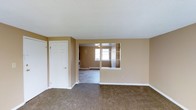 This single-floor space is designed with a functional, open layout that promotes a sense of continuity between rooms. The living area is finished with wall-to-wall carpeting in a neutral brown tone, complementing the warm beige walls that carry throughout the unit. A textured white ceiling adds a classic touch overhead.

A defining architectural feature is the built-in pass-through window with open shelving that connects the living space to the adjacent kitchen or dining area, allowing light and sightlines to flow between the two zones. This design element adds visual interest while maintaining a practical connection between spaces.

The secondary room, visible through the pass-through and an open doorway, benefits from natural light provided by a window along the far wall. A baseboard heater beneath the window suggests an efficient heating setup. The kitchen area, partially visible, includes what appears to be a white range.

A six-panel white door with a closet rod is positioned along one wall of the main living area, likely serving as a coat or storage closet. White baseboards and trim throughout provide clean contrast against the carpeting and walls, contributing to a tidy, move-in-ready aesthetic.