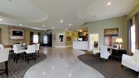 This single-floor space presents a polished and professionally designed interior that blends functionality with an inviting ambiance. The entry and main area are anchored by striking, high-gloss granite tile flooring that flows in a curved path throughout the space, creating a natural sense of movement and direction. Recessed lighting and elegantly shaped tray ceilings add architectural depth to the bright, white overhead surfaces, enhancing the overall sense of openness.

The walls are finished in a warm sage green tone, providing a calming backdrop that complements the neutral furnishings. A central reception or service desk with a clean white finish serves as the focal point of the main hall, suggesting a professional or hospitality-oriented purpose. To one side, a comfortable lounge area features white upholstered seating arranged on a patterned area rug, alongside a dark wood armoire and decorative wall art. On the opposite side, white high-back chairs are arranged around a second seating area, also set on a complementary rug. In the background, a dedicated business center alcove is visible, equipped with computer workstations. Abstract artwork and decorative wall pieces are displayed throughout, contributing to the space's refined, welcoming character.