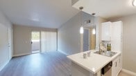This well-designed single-floor residence offers a clean, contemporary aesthetic throughout. The open-concept layout connects the kitchen and living area seamlessly, creating a spacious, airy feel enhanced by vaulted ceilings that add vertical dimension to the main living space. Light-toned hardwood-style flooring runs throughout the living and dining areas, contributing to a bright and cohesive atmosphere.

The kitchen is thoughtfully appointed with white shaker-style cabinetry, light quartz countertops, and a full suite of stainless steel appliances, including a refrigerator, range, microwave, and dishwasher. A peninsula with pendant lighting serves as both a functional workspace and a casual dining surface, anchoring the kitchen's connection to the living area. A sliding glass door in the living room provides access to an outdoor space and allows natural light to fill the interior.

The bedroom features soft carpeting and direct access to a generously sized walk-in closet equipped with built-in shelving and hanging rods, offering ample storage. The bathroom is clean and functional, featuring a full tub-shower combination with a curved curtain rod. Neutral gray tones throughout the unit create a calm, modern ambiance that is both versatile and inviting.