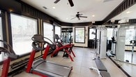 Located at 980 Courthouse Rd in Gulfport, Mississippi, this fitness center is a well-appointed, single-floor facility designed to support a comprehensive workout experience. The space features a distinctive interior design with dark wood-paneled wainscoting contrasted against light upper walls, giving the room a bold and structured aesthetic. Large windows with horizontal blinds line multiple walls, allowing abundant natural light to fill the space while offering views of the surrounding outdoor patio and landscaping.

The fitness center is equipped with a full suite of Cybex cardio machines, including treadmills and ellipticals, as well as a multi-station strength training cable machine. A dedicated free weight area features a fully stocked dumbbell rack, complemented by a variety of medicine balls and stability balls for functional training. A weight bench adds to the versatility of the strength training zone. Full-length mirrors are mounted along the walls, enhancing the sense of space and allowing users to monitor their form.

Overhead, multiple ceiling fans provide consistent air circulation throughout the facility. Recessed lighting supplements the natural light for well-lit conditions at any time of day. Durable, easy-to-clean tile flooring with rubber mat sections in the equipment areas ensures safety and comfort. A keyed entry door with secure access control provides convenient and controlled entry to the facility.