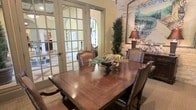 This single-story space showcases a refined Mediterranean-inspired aesthetic, combining classic architectural details with warm, welcoming interiors. The dining area serves as a focal point, centered around a substantial dark wood dining table surrounded by upholstered leather chairs, creating an atmosphere of traditional elegance. A decorative centerpiece with natural greenery adds a subtle organic touch to the space.

French doors with multi-pane glass panels open to adjacent areas, allowing natural light to filter through and creating a seamless flow between rooms. Arched entryways — including a distinctive arched door with ornate wrought iron detailing — reinforce the Mediterranean character of the design and add architectural interest throughout.

The walls feature a combination of smooth plaster in warm neutral tones and textured exposed brick or stone accents, contributing to the rustic yet refined ambiance. A large decorative painting in vibrant tones adorns one wall, adding color and artistic character to the room. A classic table lamp and a tall potted topiary further enhance the sense of curated, traditional décor. The overall atmosphere is one of timeless sophistication, with layered textures and carefully considered details that give the space a warm, livable charm.