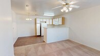 This single-floor residence offers a practical and comfortable open-concept layout that connects the living area and kitchen in one cohesive space. The kitchen is well-appointed with light wood-toned cabinetry providing generous storage, white appliances including a full-size refrigerator and microwave, and a peninsula breakfast bar that serves as both a functional workspace and a natural divider between the kitchen and living areas. The kitchen features hard surface flooring, while the living area transitions to plush wall-to-wall carpeting, adding warmth and comfort underfoot. Overhead, a ceiling fan with an integrated light fixture keeps the main living space bright and well-ventilated, while a decorative chandelier adds a touch of elegance near the dining area. The neutral color palette throughout — soft whites and warm beige tones — creates a clean, airy ambiance that lends itself well to a variety of furnishing styles. The open layout maximizes the sense of space, making the interior feel inviting and functional. Overall, the residence presents a straightforward, move-in-ready layout suited for comfortable everyday living.