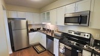 This single-story space showcases a thoughtfully designed kitchen that balances aesthetics with functionality. The kitchen features a full suite of stainless steel appliances, including a refrigerator, electric range with a built-in overhead microwave, and a dishwasher, providing everything needed for modern cooking and entertaining. White shaker-style cabinetry lines the walls from floor to ceiling, offering generous storage throughout. Upper cabinets include glass-panel accent doors, adding a touch of visual interest and elegance to the overall design. Dark granite or granite-style countertops provide a striking contrast against the bright white cabinetry, creating a classic two-tone aesthetic. The undermount sink is positioned along the countertop run, accompanied by a contemporary arc-neck faucet. Warm hardwood-style flooring runs throughout, adding depth and warmth to the space. Under-cabinet lighting enhances both the functionality and ambiance of the kitchen, ensuring well-lit prep areas. The textured ceiling and neutral wall tones contribute to a cohesive, welcoming atmosphere. Overall, the kitchen is designed to be both practical and visually appealing.