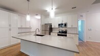 Located on Astoria Circle in Bradenton, Florida, this single-story home offers a clean, modern design throughout. The heart of the home is a well-appointed kitchen featuring crisp white shaker cabinets, a white quartz countertop island with an undermount sink, a classic subway tile backsplash, and a full suite of stainless steel appliances including a refrigerator, electric range, microwave, and dishwasher. Stylish pendant lighting hangs above the island, adding a contemporary touch. Light wood-look flooring flows through the main living areas, creating a bright and cohesive aesthetic. The open layout connects the kitchen seamlessly to the spacious living area, which benefits from natural light and a ceiling fan. A dedicated laundry area with a full-size washer and dryer is conveniently located within the home. The bedroom features plush carpeting and generous proportions, accompanied by a walk-in closet with wire shelving. An en-suite bathroom includes a wood-toned vanity and a window for natural light. The attached garage features a wide roll-up door, a concrete floor, and an electrical panel, providing practical utility and secure parking. Overall, the home presents a fresh, move-in-ready interior with functional, well-considered details throughout.