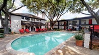 This single-floor residence is set within a community centered around a generously sized outdoor swimming pool, framed by mature shade trees and surrounded by a spacious pool deck furnished with bold red lounge chairs and market umbrellas. The two-story building exterior features a classic mid-century aesthetic with dark trim and red accent shutters, creating a striking visual backdrop.

Inside, the living area is finished with warm hardwood-style flooring, crown molding, and a ceiling fan, with windows dressed in white blinds that allow ample natural light. The open layout connects seamlessly to the kitchen, which is outfitted with dark espresso cabinetry, a tile backsplash, granite countertops, a peninsula with a stainless steel sink, pendant lighting, and black appliances. Track lighting adds functional illumination throughout.

A dedicated built-in workspace features a granite-topped desk surface, dark shelving above, and a decorative column detail—ideal for a home office setup. A conveniently located laundry closet with washer and dryer hookups is tucked just off the kitchen. Crown molding is carried throughout, lending a cohesive, refined finish to the entire space.