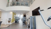 This fitness center showcases a sophisticated two-level layout, with a curved mezzanine level visible overhead, enclosed by sleek glass and stainless steel railings. The upper level is dedicated to cardio training, lined with a row of treadmills beneath ceiling fans that ensure comfortable airflow during workouts. The main floor below offers generous open space housing a comprehensive selection of Matrix strength-training machines and multi-use cable stations, thoughtfully arranged to allow easy movement between equipment. A wall-mounted flat-screen television provides entertainment for users on the main level. Large windows along the far wall flood the space with natural light, creating an inviting and energizing atmosphere. An accent wall in a muted yellow-green tone adds a pop of color, complementing the otherwise neutral white walls and light gray flooring. Recessed lighting throughout ensures consistent illumination across both levels. Ceiling fans on the main floor mirror those above, reinforcing the cohesive design. The overall ambiance is clean, modern, and functional, reflecting a well-planned facility designed to support a full range of fitness activities.
