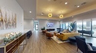 Located at 502 NW 207th St in Miami Gardens, Florida, this single-story community clubhouse is designed with a contemporary aesthetic that blends comfort and functionality throughout its thoughtfully arranged spaces. The entrance foyer makes an immediate impression with soaring ceilings, large floor-to-ceiling glass doors and transom windows that flood the space with natural light, complemented by bold orange velvet accent chairs, decorative wall art, and bamboo stalk installations flanking the entryway.

Adjacent to the entry is a dedicated Coffee Bar, set within a recessed niche with slate-blue cabinetry, marble-style countertops, and warm gold wall sconces. A coworking nook nearby features high-top wooden tables and leather-toned seating, adorned with a striking series of blue-and-white abstract art panels. The main lounge area is expansive and open, anchored by a tray ceiling with dramatic sputnik-style gold pendant fixtures. Richly toned furnishings in mustard, cognac, and navy are arranged across wood-look tile flooring, creating inviting conversation areas. A mounted flat-screen TV and a foosball table occupy the game zone, while large sliding glass doors open to views of an outdoor pool and surrounding landscape, filling the space with warmth and an airy South Florida ambiance.