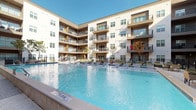 Located at 8255 Walnut Hill Ln in Dallas, Texas, this amenity space is designed to offer residents a resort-inspired social environment. At the heart of the outdoor area is a expansive, sparkling swimming pool surrounded by a generous sundeck lined with lounge chairs — ideal for relaxing under the Texas sun. The multi-story residential building wraps around the courtyard, providing a sense of enclosure and community while individual balconies overlook the pool below.

Seamlessly connected to the pool deck is a covered outdoor lounge and dining pavilion. This semi-open space features light wood-plank flooring, ceiling fans, and mounted flat-screen televisions, creating a comfortable setting for both casual gatherings and al fresco dining. Wicker lounge seating arranged over a bold blue-and-white botanical area rug anchors the social lounge area, while a tall communal dining table with rattan chairs provides a dedicated space for group meals.

Large glass sliding doors and floor-to-ceiling glazing connect the covered pavilion to an adjacent interior amenity room, flooding both spaces with natural light and maintaining visual continuity between indoors and out. White shiplap exterior cladding and clean architectural lines give the space a contemporary, coastal-inspired aesthetic that is both inviting and functional.