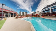 Located at 1801 E 121st Ave in Northglenn, Colorado, this resort-style pool area is a standout outdoor amenity designed for relaxation and recreation. The centerpiece is a large, rectangular swimming pool with crystal-clear blue water, complemented by an adjacent spa, providing options for both active swimming and leisurely soaking. The expansive pool deck is finished in light-toned concrete and furnished with an array of chaise lounges in bold red and navy, accented with striped and solid cushions, creating a vibrant and inviting atmosphere. Shaded seating areas are available with oversized red umbrellas, offering respite from the Colorado sun. The deck also features comfortable lounge groupings with side tables, ideal for socializing or unwinding. A covered pergola with warm wood accents provides additional shelter and a casual gathering spot. The pool area is enclosed by sleek dark-framed fencing with integrated landscaping, including ornamental grasses and young trees that add a natural touch to the setting. The surrounding multi-story residential buildings frame the space, and the open sky above allows for abundant natural light throughout the day. Signage and safety stations are thoughtfully positioned around the perimeter, ensuring a well-organized and secure environment for all users.