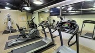 This fitness center is designed to provide a comprehensive workout environment within a compact, efficiently organized space. The room is outfitted with multiple cardio machines, including treadmills and elliptical trainers, arranged to maximize accessibility and usability. A mounted flat-screen television is positioned prominently on the wall, allowing users to stay entertained during their workout sessions. Full-length mirrors line the walls, visually expanding the space while allowing users to monitor their form. The ceiling features an industrial-style exposed design with integrated lighting that illuminates the room effectively. Additional strength and resistance equipment is visible toward the rear of the space, offering a well-rounded range of exercise options. The flooring includes protective matting beneath the equipment, ensuring safety and stability during use. The overall layout is practical and purposeful, making efficient use of the available area to deliver a functional fitness facility suited for cardiovascular training, strength work, and general exercise routines.