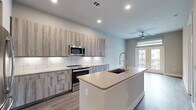 Located at 2717 Minimax Dr in Houston, TX, this thoughtfully designed one-bedroom, one-bathroom residence offers a modern living experience with high-quality finishes throughout. The open-concept layout connects the kitchen and living areas seamlessly, maximizing the efficient use of space.

The kitchen is a standout feature, equipped with stainless steel appliances including a French door refrigerator, electric range, microwave, and dishwasher. Textured wood-grain cabinetry with under-cabinet lighting pairs with a crisp white tile backsplash and light quartz countertops. A central island with an undermount sink provides additional prep space and casual dining options. Durable wood-look flooring runs throughout the main living areas.

The living room features neutral tones, a ceiling fan, and French doors that open to a private balcony, allowing natural light to fill the space. The bedroom is carpeted for comfort, features two windows with roller shades, a ceiling fan, and direct access to both a closet and the bathroom.

The bathroom is well-appointed with a dual-sink vanity, wood-grain cabinetry, quartz countertops, a large mirror, and a glass-enclosed walk-in shower with tile surround. A dedicated in-unit laundry area houses a full-size washer and dryer with overhead shelving for added storage convenience.