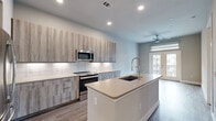 Located at 2717 Minimax Dr in Houston, TX, this thoughtfully designed one-bedroom, one-bathroom residence offers a modern living experience with high-quality finishes throughout. The open-concept layout connects the kitchen and living areas seamlessly, maximizing the efficient use of space.

The kitchen is a standout feature, equipped with stainless steel appliances including a French door refrigerator, electric range, microwave, and dishwasher. Textured wood-grain cabinetry with under-cabinet lighting pairs with a crisp white tile backsplash and light quartz countertops. A central island with an undermount sink provides additional prep space and casual dining options. Durable wood-look flooring runs throughout the main living areas.

The living room features neutral tones, a ceiling fan, and French doors that open to a private balcony, allowing natural light to fill the space. The bedroom is carpeted for comfort, features two windows with roller shades, a ceiling fan, and direct access to both a closet and the bathroom.

The bathroom is well-appointed with a dual-sink vanity, wood-grain cabinetry, quartz countertops, a large mirror, and a glass-enclosed walk-in shower with tile surround. A dedicated in-unit laundry area houses a full-size washer and dryer with overhead shelving for added storage convenience.