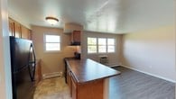 This single-floor unit is designed with practicality and comfort in mind, offering a well-organized layout across its living spaces. The kitchen is efficiently arranged with warm-toned wood cabinetry, dark granite-style countertops, and black appliances including a refrigerator, electric range, and dishwasher. A peninsula counter extends into the adjacent living and dining area, creating a natural division between the two spaces while maintaining an open feel. Tile flooring runs through the kitchen, transitioning to durable wood-look laminate flooring in the living area. The living space is bright and open, with multiple windows allowing generous natural light and views of the surrounding neighborhood. A wall-mounted HVAC unit provides climate control, complemented by baseboard heating along the perimeter walls. Two bedrooms are finished with neutral carpet and feature individual windows that bring in ample daylight, along with baseboard heating for year-round comfort. Throughout the unit, the walls are painted in soft, neutral tones, and flush-mount ceiling light fixtures provide consistent illumination. Clean white trim and baseboards complete the understated, move-in-ready aesthetic of this well-maintained space.