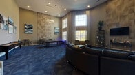 This single-floor community amenity space is designed to provide a dynamic and comfortable social environment, combining lounge and entertainment functions in one expansive, open layout. The room immediately draws attention with its soaring double-height ceilings, which amplify the sense of openness and grandeur throughout. Large, floor-to-ceiling windows dressed with patterned drapes flood the interior with natural light, offering views of the surrounding exterior.

The space is anchored by a bold, curved sectional sofa arrangement at its center, complemented by additional lounge seating including leather chairs with ottomans and accent armchairs — all set atop rich, blue geometric-patterned carpeting that ties the design together cohesively. A textured wood-panel feature wall serves as a striking architectural backdrop, paired with a wall-mounted flat-screen TV and flanked by open metal shelving units displaying curated décor.

The game area is well-appointed, featuring a full-size pool table beneath a modern geometric pendant light fixture and a shuffleboard table along the perimeter wall. Tall indoor potted plants and original artwork add warmth and a refined aesthetic throughout. The overall ambiance is modern, vibrant, and inviting — purpose-built for recreation and relaxation.