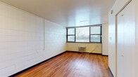 Located on Kingshighway Boulevard in St. Louis, Missouri, this single-floor unit offers a straightforward and functional layout with consistent design elements throughout. The main living area is defined by warm wood-look flooring that runs continuously through the space, white-painted concrete block walls, and multiple windows fitted with horizontal blinds that allow natural light to filter in while providing privacy. Wall-unit air conditioners are installed beneath the windows, offering climate control directly within the living space.

The open layout visible from above reveals a seamless transition between the living area and the kitchen zone. The kitchen features medium-toned wooden cabinetry, granite-style countertops, and a black ceramic glass electric range, providing a practical cooking setup. A white refrigerator and built-in ventilation are also present, completing the kitchen's functional offerings.

The bathroom is equipped with a walk-in shower stall outfitted with grab bars for added safety and accessibility, along with a handheld showerhead and standard toilet. The inclusion of accessibility features suggests the unit is designed to accommodate a range of occupants. Overall, the space presents a clean, utilitarian character well-suited for everyday living.