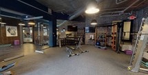 This fitness facility combines industrial-chic aesthetics with a comprehensive suite of workout equipment across a single, open floor. The design is defined by exposed brick walls, raw concrete ceilings, and visible overhead ductwork, all contributing to an urban, loft-inspired ambiance. Bold navy blue accent walls contrast with the warm tones of the brick, while pendant lighting provides focused illumination throughout the space.

The facility is divided into distinct functional zones. A dedicated strength training area features a full dumbbell rack spanning a wide range of weights, adjustable benches, a cable machine system, and an orange-accented power rack. A separate section houses plate-loaded machines and additional strength equipment positioned along the brick walls. Wall-mounted flat-screen televisions are placed throughout for entertainment during workouts.

The cardio zone is outfitted with elliptical trainers and treadmills and is partially enclosed by floor-to-ceiling glass partitions, creating a defined yet open feel. Large windows along one wall allow views to the exterior, bringing in natural light and a connection to the outdoors. Functional training accessories, including medicine balls, resistance bands, and foam mats, are neatly organized on dedicated storage racks. The flooring throughout is durable, gym-grade carpet tile suited for high-traffic use.