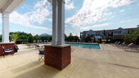 Located at 1809 Meeting St in Lexington, Kentucky, this single-level outdoor amenity space is thoughtfully designed for community recreation and relaxation. The centerpiece is a well-maintained rectangular swimming pool surrounded by a spacious concrete pool deck, complete with lounge chairs and shaded seating areas covered by umbrellas — ideal for sunny Kentucky afternoons.

Adjacent to the pool, a generously sized covered patio runs along the building's exterior and serves as the primary gathering area. The patio features classic brick columns with white-painted accents that complement the building's traditional architectural style, combining brick masonry with lap siding. A standout feature of this covered lounge area is a built-in brick outdoor fireplace topped with a wall-mounted flat-screen television, flanked by comfortable outdoor sectional sofas and a coffee table — creating an inviting entertainment hub suitable for all seasons.

The patio extends to a dedicated outdoor kitchen or bar counter area supported by matching brick pillars, offering a functional space for outdoor dining and socializing. Recycling and waste stations are conveniently integrated into the layout. Lush landscaping with ornamental trees and greenery borders the fenced perimeter, enhancing the overall ambiance with a sense of privacy and natural beauty throughout this well-appointed outdoor community space.
