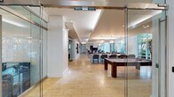 This single-floor amenity space is thoughtfully designed to offer residents a comprehensive suite of recreational and functional facilities. The layout is anchored by a spacious open lounge area bathed in natural light from expansive floor-to-ceiling windows that frame lush outdoor greenery. Tray ceilings with layered lighting and contemporary pendant fixtures — including geometric and sputnik-style chandeliers — lend an upscale, modern aesthetic throughout.

The entertainment zone features a large wall-mounted flat-screen TV with plush seating arrangements, a foosball table, a shuffleboard table, and a dedicated billiards room, all unified by warm travertine tile flooring. Adjacent to the lounge is a fully equipped catering kitchen complete with a sweeping granite island with an integrated cooktop, stainless steel appliances including a French door refrigerator and double wall ovens, and a wet bar area with a bold navy accent wall.

A business center corridor offers individual workstations along a long desk surface, complemented by stylish patterned wallcovering and glass-enclosed private meeting or conference rooms. The fitness center, enclosed in floor-to-ceiling glass, is outfitted with a full row of cardio machines and mirrored walls. The space is accessible via elevator lobbies finished in crisp white walls and travertine tile flooring.