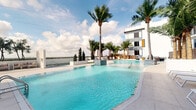This outdoor amenity space showcases a contemporary, resort-inspired design centered around a expansive swimming pool with crystal-clear turquoise water. The pool area is framed by tall, mature palm trees that add a tropical ambiance, complementing the crisp white modern architecture of the surrounding multi-story building. The structure features clean lines, large windows, private balconies with metal railings, and warm wood-toned accent panels that provide visual contrast against the white facades.

A dedicated spa or wading area connects seamlessly to the main pool, enhanced by a mosaic tile water feature that serves as a striking focal point. Along the poolside, shaded cabanas equipped with plush lounge furniture offer private retreats, while rows of white chaise lounges line the sun deck, separated from the building by horizontal wood-slat privacy fencing that adds warmth and texture to the space.

A reception or attendant desk is positioned near the entrance, framed by in-ground palm planters with black river rock detailing. Comfortable lounge seating groupings are arranged throughout the deck, ideal for socializing. The overall ambiance is bright, open, and elevated, with panoramic sky views and lush greenery contributing to an inviting, high-end outdoor living experience.