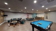 Located at 33 San Pablo Ave in San Rafael, CA, this single-floor amenity space is thoughtfully designed to serve as a vibrant social and recreational hub. The open-plan layout seamlessly connects multiple functional zones, all unified by wide-plank hardwood flooring, recessed lighting, and a contemporary color palette of warm grays and teal accents.

The game and lounge area is a standout feature, anchored by a full-size pool table with a blue felt surface and complemented by a large sectional sofa centered around a concrete coffee table atop a cowhide rug. A striking wood-paneled accent wall houses a wall-mounted flat-screen television, with a pair of teal armchairs nearby offering an additional relaxed seating nook. A mural depicting the Golden Gate Bridge adds a distinct Bay Area character to the space.

A dedicated dining area features a sturdy communal table with seating for several guests, positioned centrally within the room for easy access. Adjacent to this is a well-appointed kitchenette, outfitted with light wood cabinetry, white quartz countertops, a textured tile backsplash with under-cabinet lighting, stainless steel appliances including a French door refrigerator, wine cooler, dishwasher, and a sink. A teal accent wall and pendant lighting complete the kitchen's modern aesthetic. Restroom facilities are conveniently accessible from the kitchen corridor.