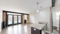Located at 310 W Sixth St in Royal Oak, Michigan, this single-floor unit offers a thoughtfully designed open-concept layout that blends industrial character with modern updates. The combined kitchen and living area serves as the heart of the space, anchored by a sleek kitchen peninsula with granite countertops, pendant lighting, and stainless steel appliances including a range and refrigerator. White upper cabinetry and a wall-mounted mini-split unit add both function and a contemporary touch. Light wood-tone flooring flows throughout, enhancing the sense of openness. The living area is defined by striking exposed brick walls and bold black-framed French doors that flood the room with natural light and offer views of the surrounding streetscape. A classic cast-iron radiator and a ceiling fan add to the unit's distinctive industrial-residential character. A mirrored closet and additional window in a secondary area provide storage and continued natural light. The bathroom features a walk-in shower with large-format dark tile walls and white mosaic tile flooring, offering a clean, modern aesthetic. Overall, this space delivers an efficient yet characterful urban living experience in the heart of Royal Oak.
