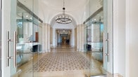 Located at 2121 S Hiawassee Rd in Orlando, Florida, this distinguished single-floor commercial building lobby showcases a seamless blend of classical and contemporary design. The grand entrance corridor immediately impresses with soaring arched ceilings, substantial travertine columns, and an intricate mosaic-patterned tile floor that runs the length of the hallway, drawing the eye deep into the space. Elegant chandelier lighting punctuates the corridor at regular intervals, casting a warm, refined glow throughout.

The lobby features a reception desk framed by ornate travertine columns, providing a formal welcome point for visitors. Floor-to-ceiling glass partition walls line both sides of the main corridor, enclosing modern office and meeting areas while maintaining an open, transparent feel. One glass-enclosed suite is furnished as a contemporary lounge with curved white sofas and sleek accent lamps, while another serves as a fully equipped workspace.

Stainless steel elevators with travertine surrounds offer seamless vertical access. Decorative touches — including a large grid-framed mirror above a fluted sideboard, ornamental urns with tropical plantings, and arched doorways — add warmth and sophistication. The main entrance features full-height glass doors that invite abundant natural light and offer views of the surrounding outdoor landscape, creating a bright and welcoming atmosphere throughout.