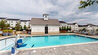 Located in Spartanburg, South Carolina, this single-story pool house serves as a functional amenity facility within a well-appointed residential community. The building's exterior showcases classic farmhouse-inspired architecture, featuring white board-and-batten siding, a standing-seam metal roof, and a charming cupola detail—complementing the larger white multi-story residential buildings visible in the background.

The structure opens directly onto a generously sized outdoor swimming pool, surrounded by a paved stone deck with seating and grilling areas nearby. A glass-paneled black entry door provides a clear view of the pool from inside, reinforcing the connection between the interior and outdoor spaces.

Inside, the facility maintains a clean, modern aesthetic with white walls, dark tile flooring, and bright natural light streaming through double-hung windows overlooking the pool area and surrounding greenery. A dual-level stainless steel water fountain station with a built-in bottle-filling option is conveniently positioned near the entrance. Separate ADA-accessible restrooms are clearly marked, with the women's restroom featuring classic white subway tile walls and a decorative patterned tile floor. The interior also includes a wall-mounted HVAC unit, ensuring year-round comfort for guests using the amenity space.
