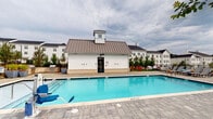 Located in Spartanburg, South Carolina, this single-story pool house serves as a functional amenity facility within a well-appointed residential community. The building's exterior showcases classic farmhouse-inspired architecture, featuring white board-and-batten siding, a standing-seam metal roof, and a charming cupola detail—complementing the larger white multi-story residential buildings visible in the background.

The structure opens directly onto a generously sized outdoor swimming pool, surrounded by a paved stone deck with seating and grilling areas nearby. A glass-paneled black entry door provides a clear view of the pool from inside, reinforcing the connection between the interior and outdoor spaces.

Inside, the facility maintains a clean, modern aesthetic with white walls, dark tile flooring, and bright natural light streaming through double-hung windows overlooking the pool area and surrounding greenery. A dual-level stainless steel water fountain station with a built-in bottle-filling option is conveniently positioned near the entrance. Separate ADA-accessible restrooms are clearly marked, with the women's restroom featuring classic white subway tile walls and a decorative patterned tile floor. The interior also includes a wall-mounted HVAC unit, ensuring year-round comfort for guests using the amenity space.