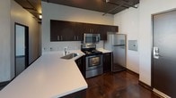 This single-floor unit showcases a distinct industrial-modern aesthetic, defined by rich, polished concrete floors that flow consistently throughout the space, creating a cohesive and striking visual foundation. The exposed ceiling infrastructure, complete with visible ductwork and track lighting, reinforces the urban loft character of the unit.

The kitchen is a functional highlight, outfitted with dark espresso cabinetry, white countertops, and a full suite of stainless steel appliances including a refrigerator, electric range, and built-in microwave. A peninsula counter provides additional prep space and adds to the open feel of the area.

The living and bedroom areas feature clean white walls that contrast beautifully with the dark flooring, offering a versatile, gallery-like atmosphere. Ceiling fans and a wall-mounted mini-split HVAC unit provide year-round climate control, while a window allows for natural light to filter into the space.

The bathroom is finished with a sleek, floating white vanity, a large wall mirror, industrial-style lighting fixtures, and a full-size soaking tub with shower. Chrome hardware and white fixtures contribute to its clean, modern presentation. The polished concrete flooring extends into the bathroom, maintaining design continuity throughout the unit. An in-unit washer and dryer connection is also available.
