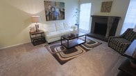 This single-floor residential space offers a comfortable and stylishly designed interior that balances modern aesthetics with a warm, inviting ambiance. The living area serves as the centerpiece of the home, featuring plush wall-to-wall carpeting that adds softness and warmth underfoot. A sleek, dark-mantled fireplace anchors one side of the room, providing a refined focal point and a cozy atmosphere. Natural light filters in through a window fitted with horizontal blinds, brightening the space and creating an airy feel. The living room is thoughtfully furnished with a tufted white leather sofa, a dark wood coffee table with a lower shelf, and a coordinating side table topped with a stylish lamp. A bold geometric area rug in earth tones anchors the seating arrangement, adding visual interest and depth. A large, vibrant abstract canvas on the wall introduces a splash of color and artistic character to the room. A potted plant in the corner contributes a touch of greenery and life to the overall setting. The combination of neutral wall tones, contemporary furnishings, and carefully chosen accents creates a cohesive, polished living environment that feels both functional and welcoming.