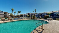 Located at 20251 N 75th Ave in Glendale, Arizona, this single-level outdoor amenity area showcases a generously sized resort-style swimming pool as its centerpiece. The pool features crystal-clear turquoise water, dual stainless steel entry rails, and a broad surrounding deck finished in textured concrete, providing ample space for relaxation and recreation. A dedicated hot tub spa is situated adjacent to the main pool, complete with its own entry rail and tiled interior, offering a comfortable retreat. The pool deck is well-appointed with multiple lounge chairs and seating areas, including a shaded covered patio with outdoor furniture, ideal for escaping the Arizona sun. Lush, manicured landscaping borders the area, featuring tall palm trees, flowering shrubs, and trimmed hedgerows that add vibrant color and privacy. A secure wrought iron fence with gated entry surrounds the entire pool area. In the background, the multi-story residential buildings present a classic Southwestern architectural style with neutral stucco exteriors, tiled rooflines, and balconies. The community clubhouse, visible from the pool, features large windows, arched architectural accents, and stone detailing. The space enjoys abundant natural light and open sky views year-round, characteristic of the sunny Glendale, AZ climate.