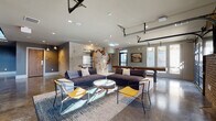 Located at 1835 Morehead Ridge Dr in Charlotte, NC, this contemporary community clubhouse showcases a bold industrial aesthetic defined by polished concrete floors, exposed structural elements, and soaring ceilings. The open-plan layout flows seamlessly between distinct zones, creating a versatile social environment. A standout lounge area anchors the space with deep sectional sofas, a marble-topped coffee table, and vibrant yellow accent chairs arranged around a woven area rug. The focal point of this seating area is a dramatic concrete feature wall adorned with a vivid koi fish mural, adding an artistic, urban character to the room. A dedicated game area includes a ping pong table, while a shuffleboard table offers additional recreation nearby. Industrial-style garage doors open the space to the outdoors, flooding the interior with natural light and providing views of the surrounding greenery. A bar-height counter with sleek black stools runs along a wall of floor-to-ceiling windows, offering additional seating bathed in natural light. A steel staircase with cable railings leads to an upper level, anchored by lush built-in planters that introduce organic warmth to the industrial palette. Recessed lighting, track lighting, and strategically placed pendant fixtures ensure a well-lit and inviting ambiance throughout.
