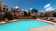 This amenity space features two distinct areas designed to support an active lifestyle. The outdoor pool area showcases a generously sized, turquoise swimming pool surrounded by a concrete deck with lounge seating. A black wrought-iron fence encloses the pool area, complemented by neatly trimmed hedgerows, ornamental plantings, and mature trees that provide shade and a park-like atmosphere. The surrounding residential buildings, featuring classic architectural details such as pitched rooflines, chimneys, and arched windows, frame the space with a cohesive aesthetic. Patio umbrellas and additional seating areas are visible nearby.

The fitness center is a spacious, single-level facility filled with abundant natural light from multiple large windows fitted with white blinds, offering views of the community grounds and pool area. The room features high ceilings with ceiling fans and recessed lighting, along with herringbone-patterned carpet flooring throughout. Equipment includes a multi-station functional training rig, a full dumbbell rack, medicine balls, resistance battle ropes, a cable machine, a power rack with bench, a rowing machine, a stationary bike, and treadmills. A wall of mirrors visually expands the space, while a wall-mounted television enhances the cardio area.