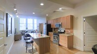 This single-floor space is designed with an open-concept layout that integrates the kitchen and living area into one cohesive, functional environment. The kitchen is a central highlight, outfitted with warm honey-toned wood cabinetry that extends to the upper walls, providing generous storage throughout. Stainless steel appliances, including a gas range with oven, over-the-range microwave, and a full-size refrigerator, offer modern convenience. A decorative tile backsplash adds a subtle textural accent behind the range. The kitchen island features a light granite countertop and accommodates seating with bar-height stools, making it ideal for casual dining or entertaining. Light hardwood flooring runs throughout, contributing to the warm, inviting ambiance of the space. Recessed lighting in the ceiling ensures consistent, even illumination. The open living area beyond the kitchen benefits from large windows that allow abundant natural light to fill the space, creating a bright and airy atmosphere. The overall design balances practicality with a clean, contemporary aesthetic throughout.