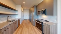 Located at 1390 NE Compton Dr in Hillsboro, Oregon, this efficiently designed one-bedroom, one-bathroom unit offers a thoughtful layout across a single floor. Light wood-tone flooring runs continuously throughout, creating a cohesive and bright aesthetic.

The open-concept living area seamlessly connects the kitchen, dining, and living spaces. The modern kitchen is well-appointed with stainless steel appliances including a refrigerator, range, microwave, and dishwasher. Sleek gray cabinetry with warm wood accents is complemented by a white subway tile backsplash and open shelving. Recessed lighting illuminates the space, while a window at the far end of the living area draws in natural light and offers outdoor views.

The bedroom is a comfortable, serene retreat with clean white walls and a large window that floods the room with natural light and provides views of the surrounding landscape. A sliding barn-style door leads to a walk-in closet outfitted with wire shelving and hanging rods, providing ample storage. An additional closet offers further organizational flexibility.

The bathroom and laundry room are combined into one practical space, featuring light wood flooring, a wood-toned vanity, and a clean, minimalist design with grab bar accessibility features. The unit's modern, no-fuss aesthetic and functional layout make it a practical and attractive living space.