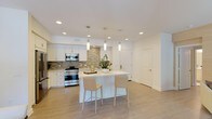 This single-floor space showcases a thoughtfully designed open-concept layout that seamlessly blends style and functionality. The kitchen is a standout centerpiece, featuring crisp white cabinetry, a textured mosaic tile backsplash, and premium stainless steel appliances including a professional-grade range and built-in microwave. A sleek white kitchen island serves as a natural gathering point, accommodating bar-style seating with modern cream-toned chairs — ideal for casual dining or entertaining. Elegant cylindrical pendant lights hang above the island, complementing the recessed lighting throughout the space to create a bright, welcoming atmosphere. Light-toned tile flooring extends seamlessly across the open area, enhancing the sense of spaciousness and contributing to the clean, contemporary aesthetic. The open layout connects the kitchen to adjacent living areas, allowing natural flow and versatility in how the space can be used. White walls and a neutral color palette throughout provide a timeless backdrop, while subtle decorative touches like potted greenery add a refreshing, organic element. Overall, the space offers a polished, modern environment that balances clean architectural lines with warm, livable design.