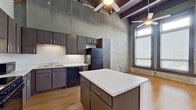 Unit 206 is a single-floor loft-style space that showcases an impressive blend of industrial heritage and contemporary design. The defining architectural feature is the dramatically tall ceiling, accentuated by dark exposed wooden beams and ceiling fans that draw the eye upward, creating a spacious, open atmosphere throughout.

The kitchen serves as a central hub, offering an abundance of dark cabinetry extending nearly to the ceiling, white countertops, and a full suite of black appliances including a range, dishwasher, microwave, and refrigerator. A generously sized center island provides additional workspace and storage, making the kitchen highly functional.

The living area retains strong industrial character through exposed concrete and partially revealed brick walls, lending authentic texture and visual depth. Warm hardwood-style flooring runs throughout, providing contrast to the raw wall surfaces. Multiple large windows with metal-framed surrounds allow natural light to fill the space, with views outward adding an airy quality.

The bathroom features a dark wood vanity with ample storage, a framed mirror, and consistent hardwood flooring that maintains the cohesive aesthetic found throughout the unit. Overall, Unit 206 delivers a distinctive urban loft ambiance defined by its raw materials, vertical scale, and modern conveniences.