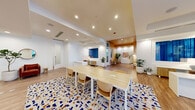 This thoughtfully designed single-floor commercial space makes a strong first impression with a sleek glass-enclosed lobby entrance, framed by a graceful arched ceiling and warm wood-look flooring that flows throughout the entire space. The grand glass entry doors invite abundant natural light and offer clear sightlines to the exterior.

Inside, the open layout transitions seamlessly between distinct functional zones. A central collaborative area features a generously sized light wood conference table surrounded by curved natural-wood chairs, anchored by a bold terrazzo-style area rug in blue, white, and amber tones that adds a striking design focal point. Recessed lighting illuminates the space evenly overhead, complemented by warm accent zones with golden-hued ceilings.

A more relaxed lounge area incorporates rattan rocking chairs and woven pendant lights near floor-to-ceiling windows draped with rich navy curtains, creating a casual yet stylish retreat. An adjacent nook showcases a white-painted brick accent wall adorned with contemporary artwork, a credenza, and lush potted greenery.

Private office areas are accessible via glass-paneled sliding doors, maintaining visual openness while providing separation. A dedicated workstation with white cabinetry and a planter adds functional elegance. Curated artwork, abundant indoor plants, and cohesive warm-toned furnishings throughout contribute to an inviting, creative, and professional ambiance.