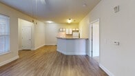 Located at 202 Ashbury Square in Mebane, North Carolina, this single-floor residence offers a practical and well-appointed layout designed for comfortable everyday living. The open-concept living and kitchen area serves as the heart of the home, with warm hardwood-style flooring flowing throughout the main spaces. The living room is bright and spacious, featuring a ceiling fan with light fixture and multiple windows dressed with blinds that invite natural light while maintaining privacy. A peninsula counter with a built-in sink separates the kitchen from the living area, creating a functional and social layout. The kitchen is equipped with a range, refrigerator, and ample upper cabinetry in a warm wood-tone finish, complemented by overhead lighting. Carpeted bedrooms provide a softer, quieter retreat, with neutral wall tones and windows offering views of the surrounding greenery. The bathroom is thoughtfully designed with accessibility in mind, featuring stainless steel grab bars around the tub-shower combination, a wall-mounted sink, a medicine cabinet with mirror, and hardwood-style flooring. Clean lines, neutral color palettes, and consistent finishes throughout give the home a cohesive and understated aesthetic well-suited for a range of occupants.