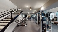 This expansive two-floor fitness center is thoughtfully designed to accommodate a full range of workout needs. The lower level features durable rubber flooring and houses an extensive strength training area equipped with cable machines, including the Technogym Instinct system, a comprehensive free weight section with dumbbells, benches, and weight racks, and a selection of functional training accessories. A built-in storage unit keeps the space organized, and recessed lighting maintains a clean, focused atmosphere. A bold open staircase with metal railings connects the two levels, offering an architectural focal point throughout the facility. The upper floor transitions to light wood-toned flooring and is dedicated to cardiovascular training, lined with a row of Star Trac treadmills and elliptical machines along mirrored and windowed walls that invite natural light. At the far end of the upper level, a separate cycling studio provides an immersive workout environment, featuring spin bikes, a large wall-mounted display, medicine ball racks, battle ropes, and surround sound speakers. A tray ceiling with perimeter lighting adds a polished finish to this studio space. Throughout both floors, wall-mounted televisions, sanitation stations, and ample open floor space reflect a facility designed with both performance and convenience in mind.