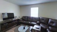 Located at 160 Broadway St in Clinton, Mississippi, this single-story home offers a comfortable and functional living environment. The living room serves as the heart of the space, featuring a large dark brown leather sectional sofa that anchors the room with a sense of warmth and ease. A textured ceiling adds subtle architectural character overhead, while neutral-toned walls create a clean, understated backdrop throughout. Natural light filters in through a window fitted with horizontal blinds, brightening the room and contributing to its welcoming atmosphere. The living area is thoughtfully arranged with a glass-top coffee table and a coordinating side table, providing practical surface space. A dark wood media console houses a flat-screen television, completing the entertainment setup. Carpeted flooring adds softness underfoot and enhances the cozy feel of the room. Personal touches, including framed photos displayed on the media console, lend a lived-in, homey quality to the space. Overall, this home presents a straightforward, comfortable layout well-suited for everyday living in a quiet Clinton neighborhood.