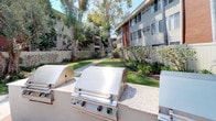This residential community presents a welcoming exterior characterized by a mid-century-influenced multi-story building with a distinctive two-tone facade of red and gray panels. The grounds are thoughtfully landscaped, featuring a manicured lawn, flowering shrubs, and a mix of mature palm trees and broad canopy shade trees that provide a sense of established greenery throughout the property.

A central courtyard walkway connects the buildings, lined with well-tended garden beds filled with tropical and ornamental plantings. The pathway creates a pleasant pedestrian corridor between structures, enhancing the sense of community within the complex.

A dedicated outdoor grilling area serves as a notable communal amenity, equipped with multiple stainless steel built-in BBQ grills mounted on a concrete station. Seating nearby allows residents to comfortably enjoy the outdoor space. Vinyl fencing borders portions of the property, offering privacy while maintaining an open, airy feel.

Upper-level units feature balconies that overlook the courtyard below, adding an outdoor extension to interior living spaces. The overall ambiance is calm and well-kept, with abundant natural light and mature landscaping contributing to a comfortable, inviting atmosphere.